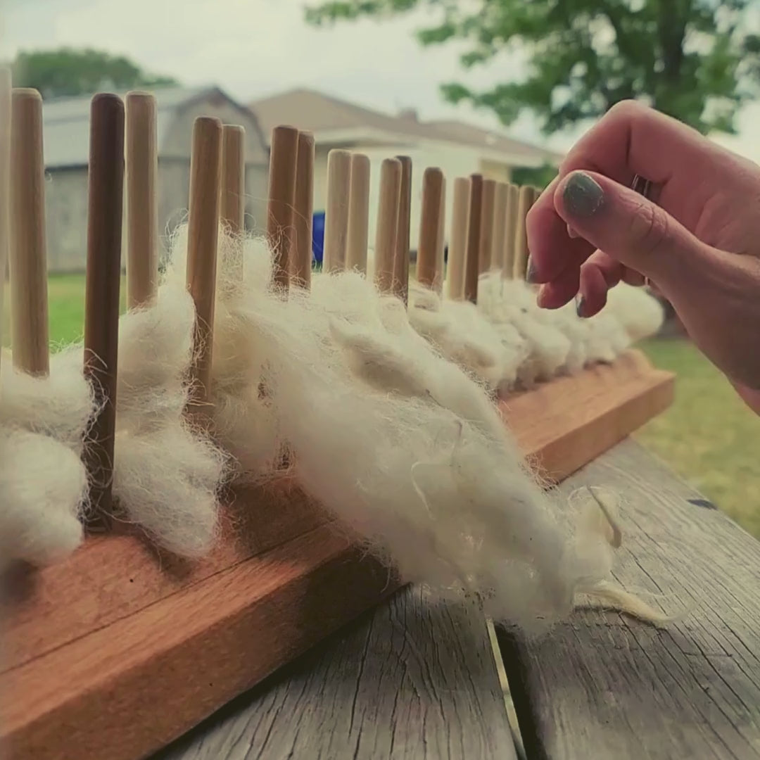 Load video: The artist sitting outside at a picnic table weaving on a peg loom, creating a rug using white sheep wool.